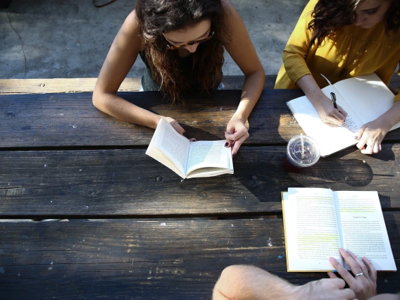 A group of people sitting on a wooden table during the day and writing in their notebooks, drinking coffee and talking to each other as the sun shines on their faces